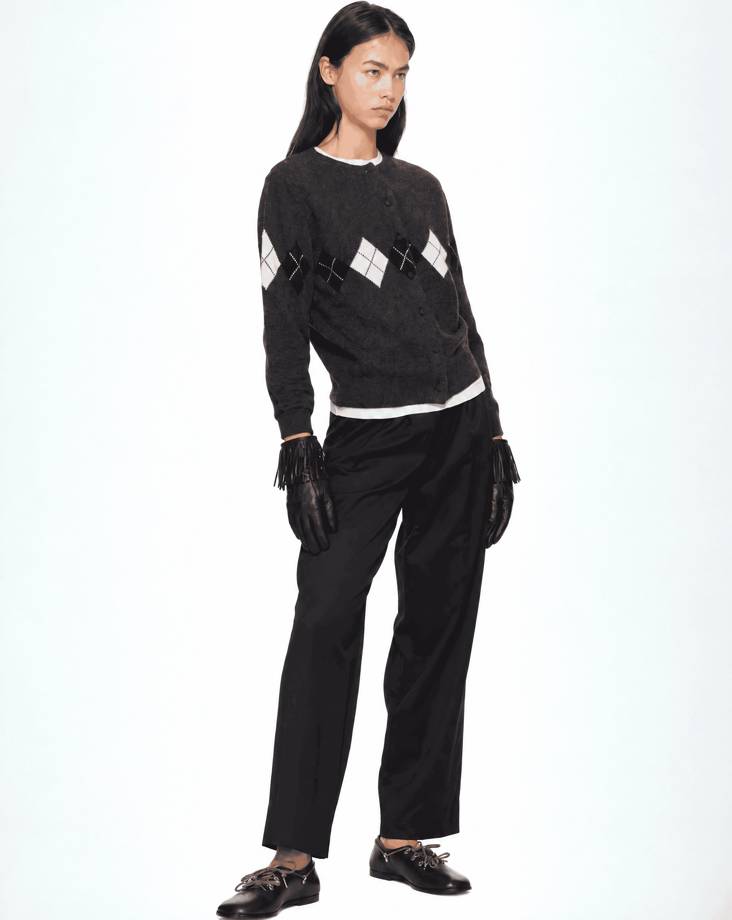 a woman in a charcoal argyle cardigan over a white t-shirt, black trousers and black loafers, wearing black leather gloves with a long fringe at the wrist
